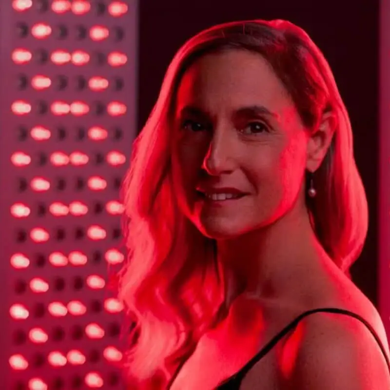 A mature woman smiling next to a red light therapy device.